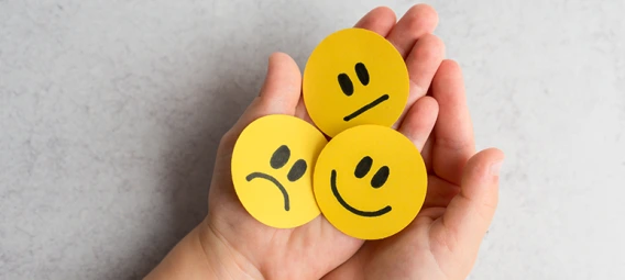 Hand holding three yellow emoji faces showing sad, neutral, and happy expressions, representing different emotional states or mood tracking