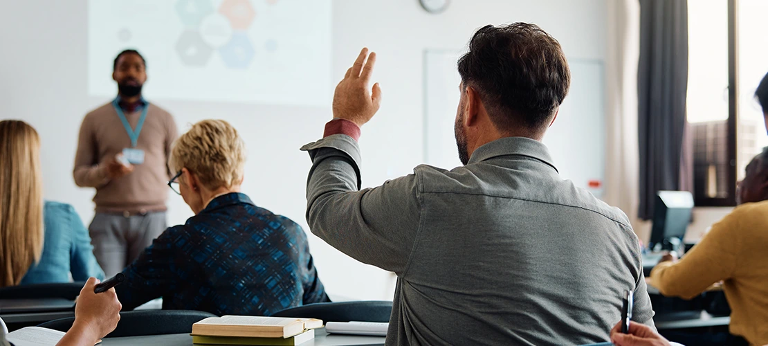 Professional training session with instructor presenting to audience, participant raising hand to ask question in modern classroom setting