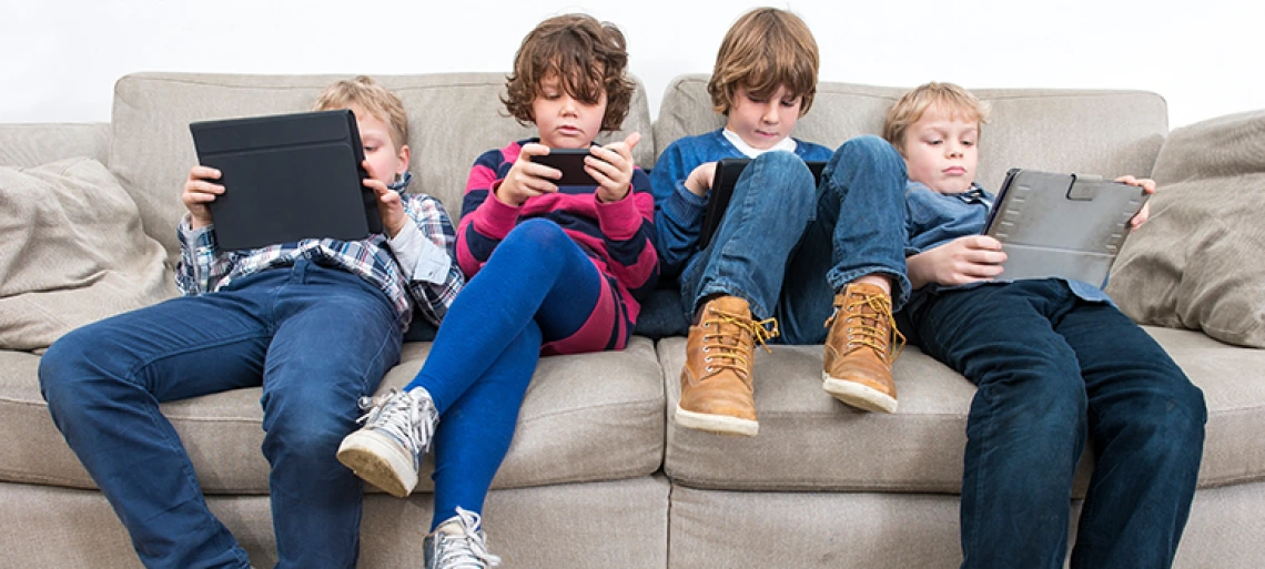 Three children sitting on gray couch using tablets and phones, representing digital device usage and screen time among young people