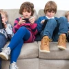 Three children sitting on gray couch using tablets and phones, representing digital device usage and screen time among young people