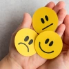 Hand holding three yellow emoji faces showing sad, neutral, and happy expressions, representing different emotional states or mood tracking